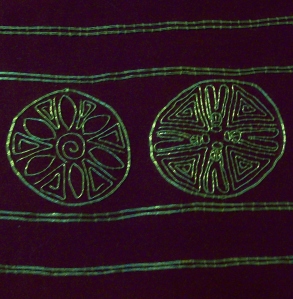 A close-up of the repeating motifs of the cuffs design; A flower in one roundel (left) and 4 bees in the other (right).